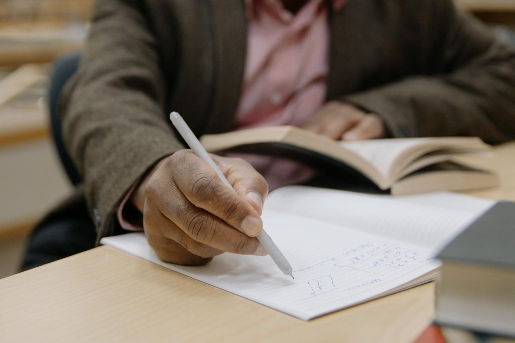 A person writing notes with a pen while studying an open book in a library setting.